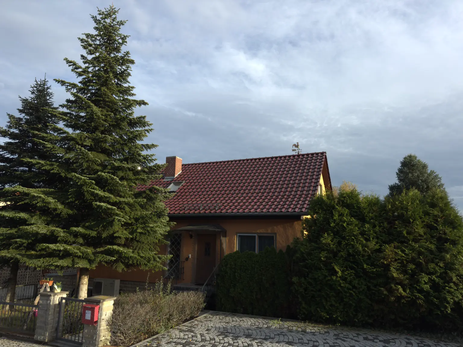Gemütliches Haus mit rotem Ziegeldach, umgeben von hohen Nadelbäumen und grünen Büschen, unter einem bewölkten Himmel in Bad Klosterlausnitz.